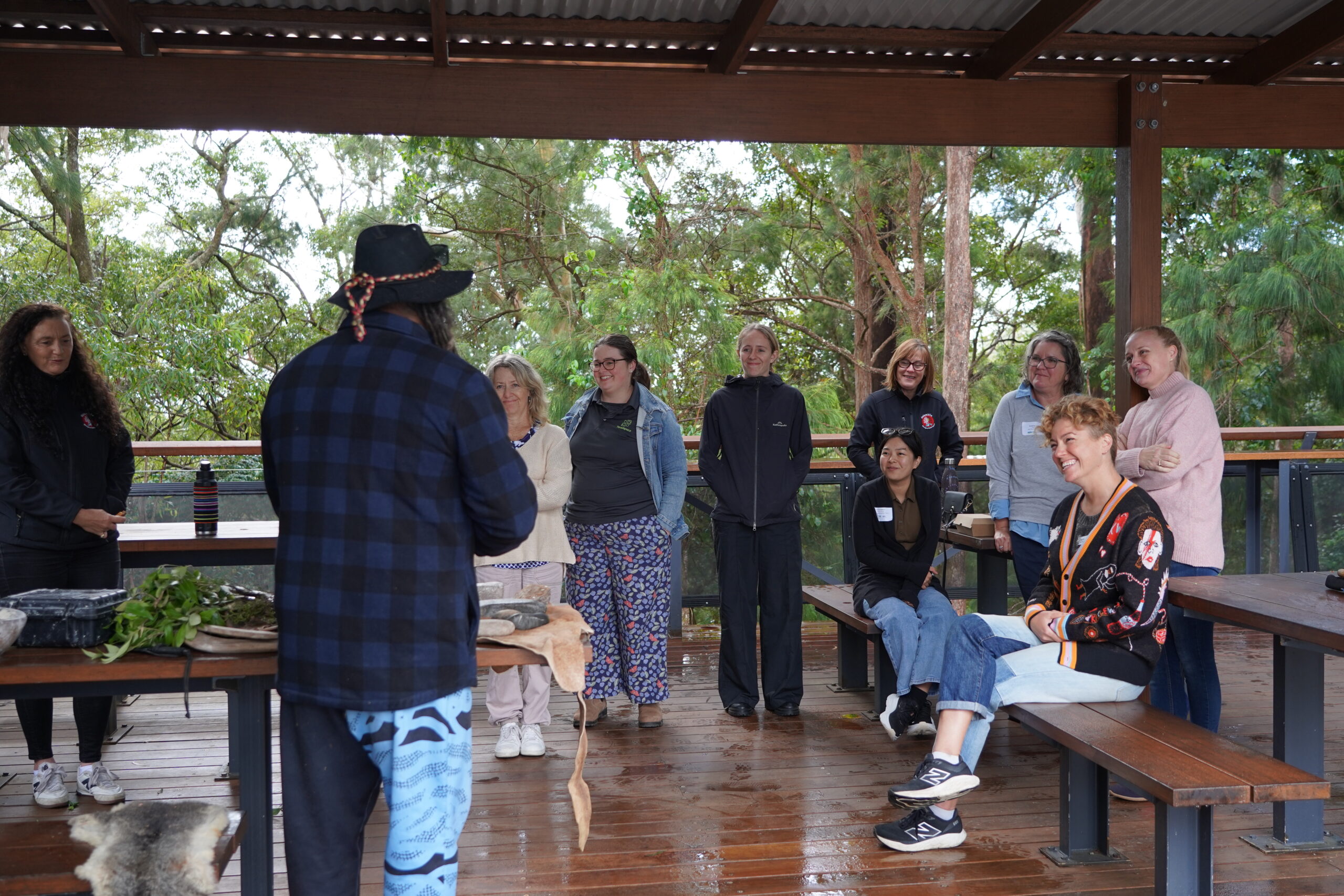 Facilitator wearing a hat speaks to a group of training participants under a covered outdoor deck surrounded by bushland, with traditional tools and native plants laid out on a table