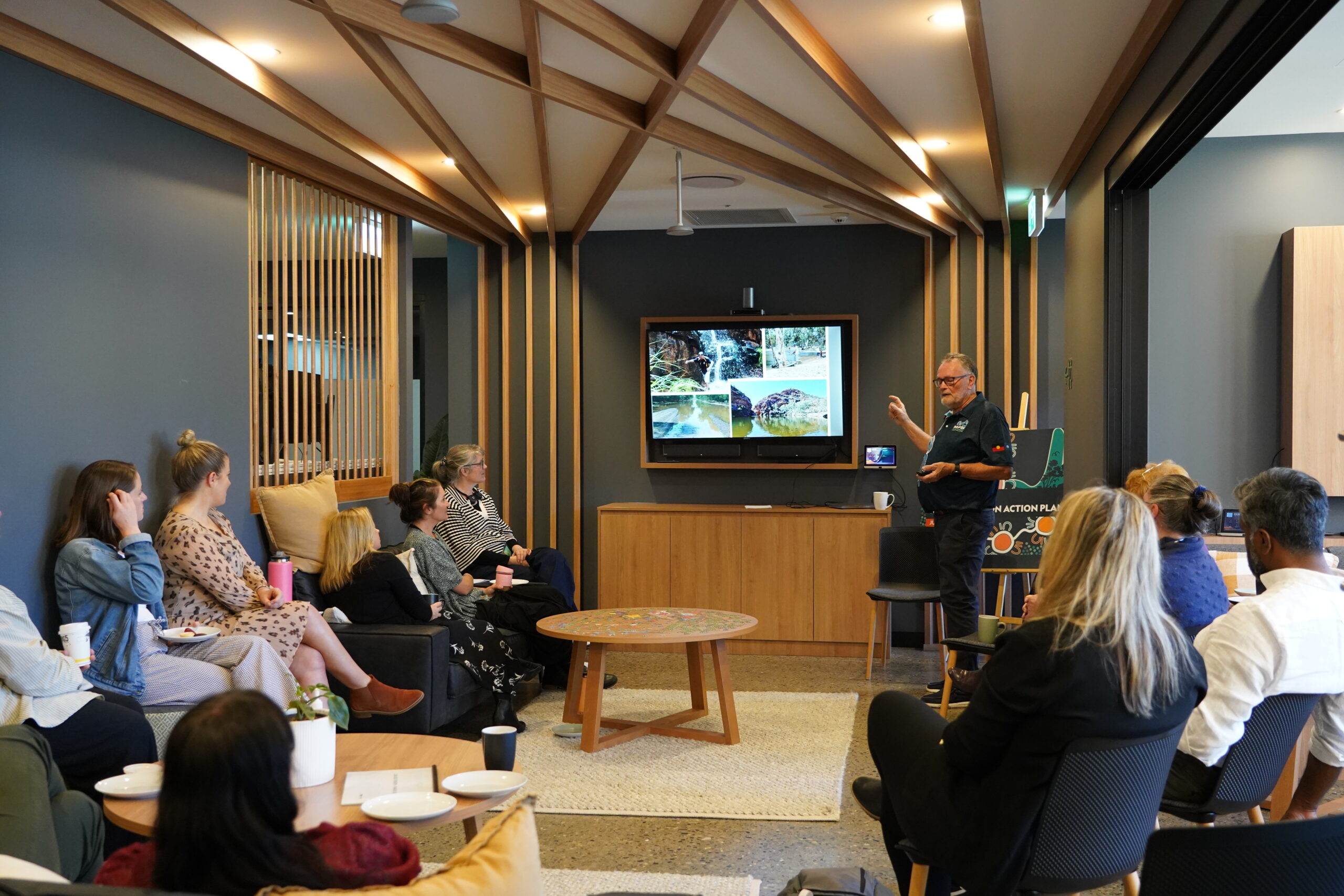 Presenter speaking to seated participants in a meeting room, with photos of waterways on a screen and an Aboriginal artwork displayed beside him