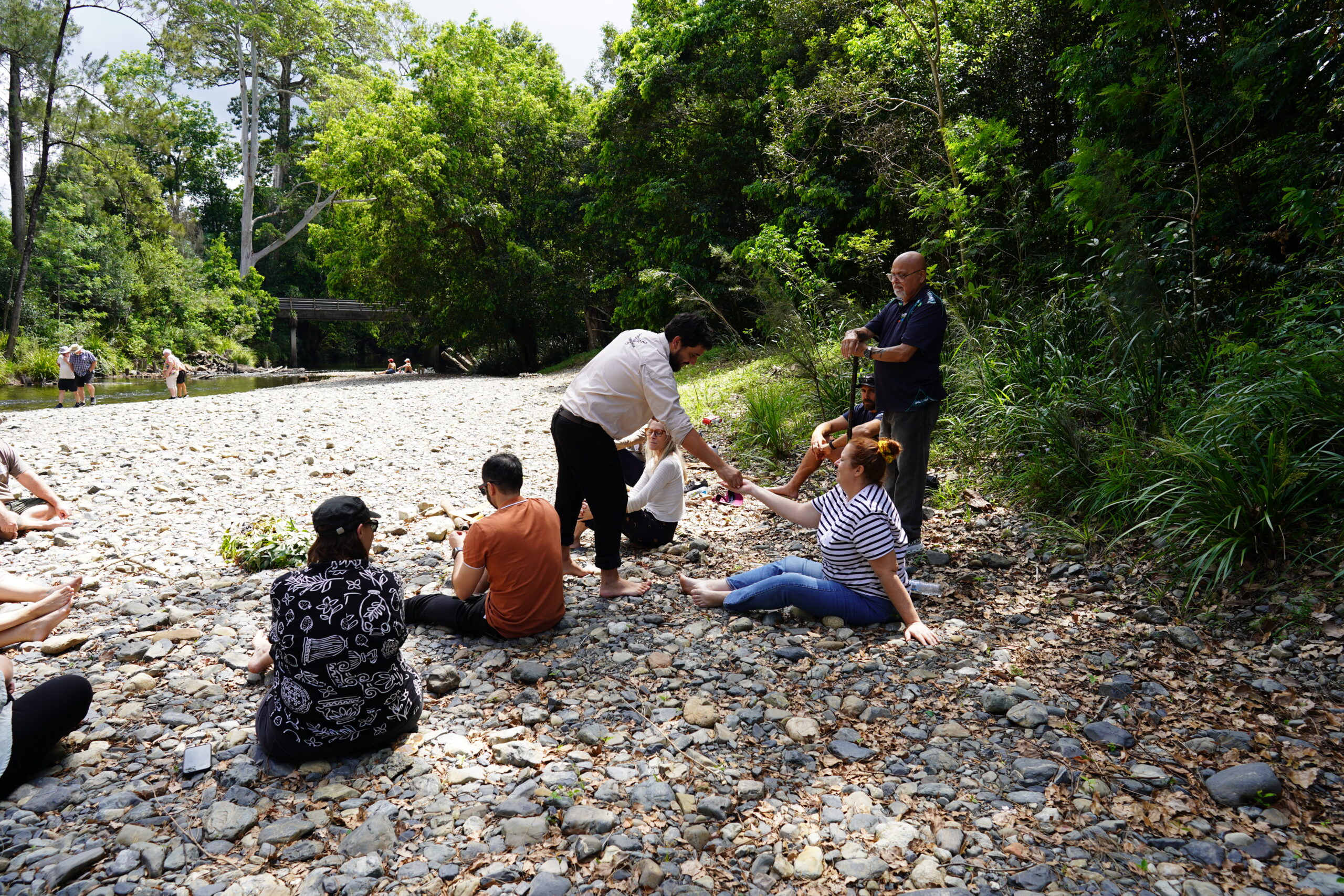 Participants seated in a circle on a pebbled riverbank during an on-Country learning session, with facilitators moving among the group