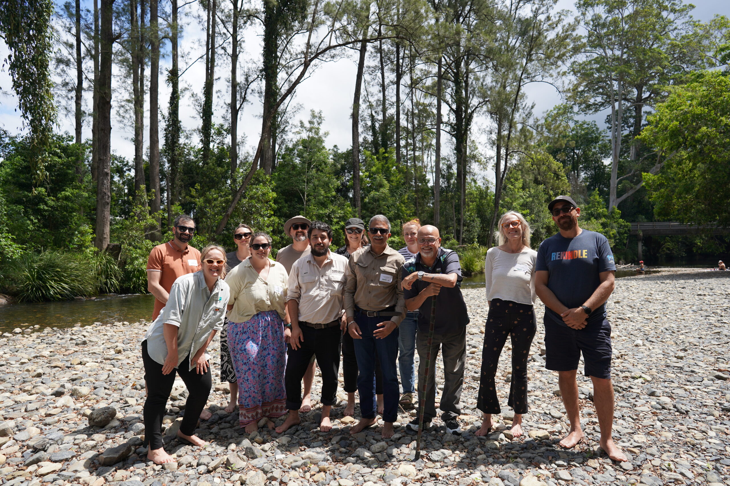 Training participants and facilitators standing together on a rocky riverbank surrounded by tall eucalypts, many barefoot after an on-Country session